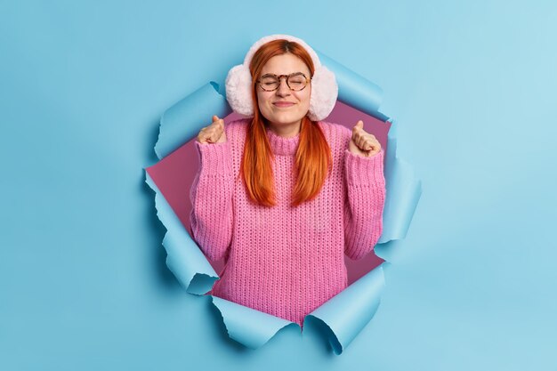 Bonita mujer pelirroja esperanzada sostiene los puños apretados y anticipa que suceda algo muy bueno hace que el deseo use orejeras de invierno y el jersey de punto se rompe a través del papel rasgado