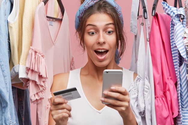 Bonita mujer de pie cerca del armario con ropa, sosteniendo un teléfono inteligente y una tarjeta de plástico, haciendo compras en línea, mirando con sorpresa en el teléfono celular mientras tiene una variedad de vestidos para elegir