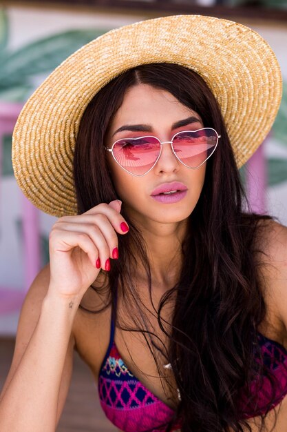 Bonita mujer de playa con traje de baño de colores brillantes, gafas de sol de corazón rosa y sombrero de paja disfrutando del verano