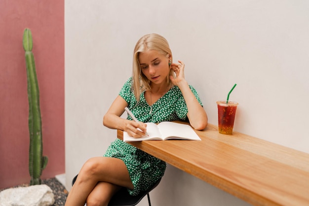 Bonita mujer rubia escribiendo en un cuaderno con vestido de verano sentada en un café moderno con paredes blancas