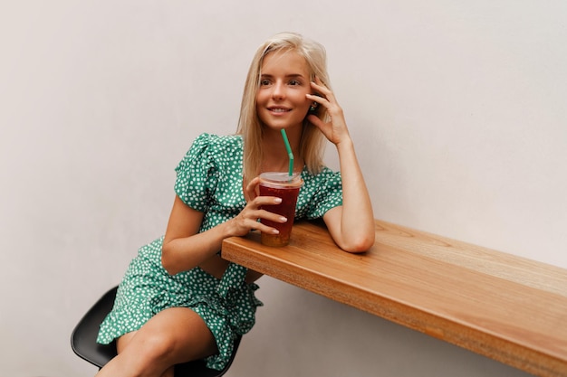 Bonita mujer rubia sentada en un café moderno y bebiendo limonada dulce con vestido elegante