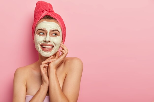 Bonita mujer sonriente con máscara de arcilla, hace un paso de belleza, limpia la cara, usa una toalla envuelta en la cabeza, se para sin camisa, obtiene placer, reduce las espinillas, copia el área del espacio contra la pared rosa