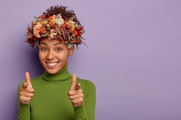 Bonita niña afroamericana apunta directamente a la cámara, hace un gesto de pistola con el dedo, sonríe positivamente, expresa su elección, usa una corona de plantas de otoño natural, vestida con un jersey informal, posa en interiores