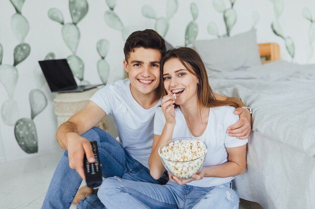 Bonita pareja joven en ropa casual viendo la televisión en casa en la cabecera y comiendo palomitas de maíz