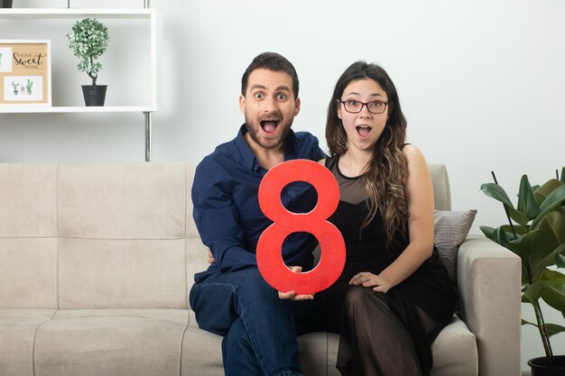 Bonita pareja sorprendida sosteniendo la figura roja de ocho sentados en el sofá en la sala de estar en el día internacional de la mujer de marzo