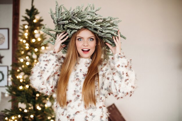 Bonita pelirroja con hermosa corona de Navidad en la cabeza y mirando a cámara con sorpresa.
