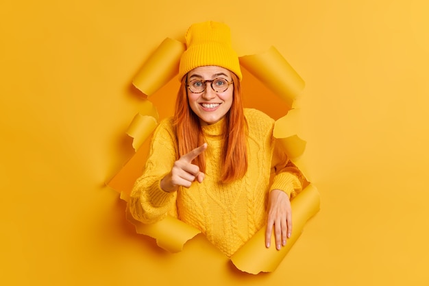 Bonita pelirroja hermosa mujer europea señala con el dedo índice directamente elige a alguien que ve algo interesante en el frente sonríe agradablemente usa sombrero suéter.