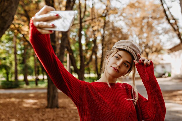 Bonita rubia haciendo selfie en el parque de otoño. Encantadora dama con suéter rojo y sombrero blanco hace fotos.