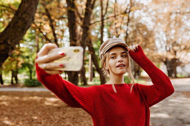 Bonita rubia haciendo selfie en el parque de otoño. Encantadora dama con suéter rojo y sombrero blanco hace fotos.