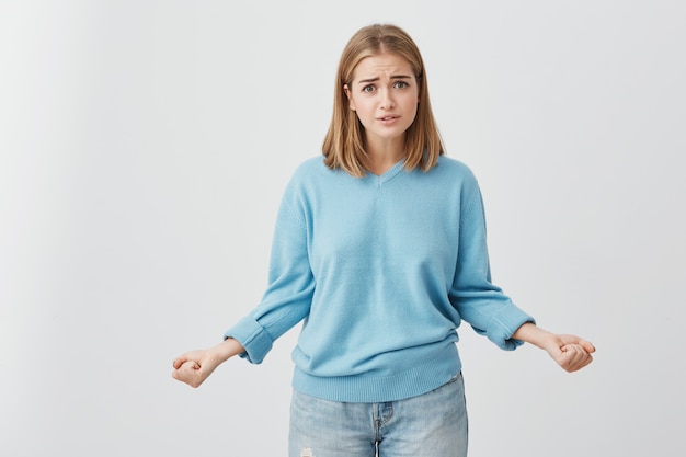 Bonita rubia joven con ojos oscuros, frunciendo el ceño mientras mira a la cámara con una mirada molesta y pensativa. Chica linda desconcertada e insatisfecha en suéter azul y jeans