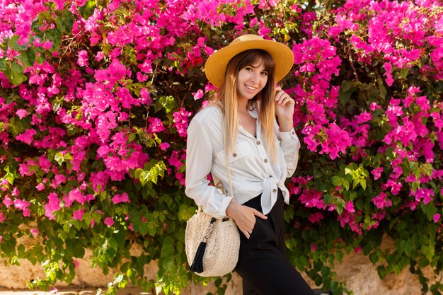 Bonita rubia mujer con sombrero de paja posando sobre rosa árbol floreciente en día soleado de primavera