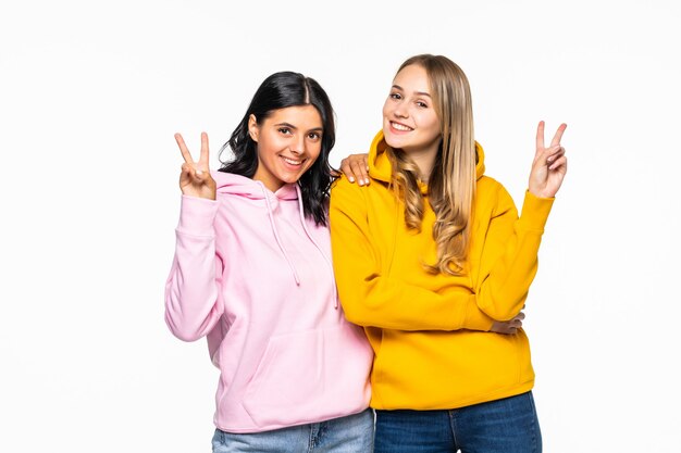 Bonitas mujeres de dos novias que muestran símbolos de signo v, vistiendo sudaderas con capucha brillantes casuales y jeans aislados pared blanca