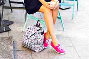 Foto gratuita bonitas piernas de mujer, niña sentada en un café al aire libre, sosteniendo una taza de capuchino, café, tarde en las manos. el uso de gumshoes rosa, elegante mochila plateada junto a los zapatos.