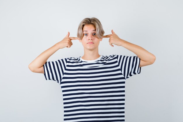Bonito muchacho adolescente tapando los oídos con los dedos en camiseta rayada y mirando desconcertado, vista frontal.