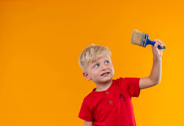 Un bonito niño con cabello rubio y ojos azules vistiendo camiseta roja levantando pincel azul mirándolo en una pared amarilla