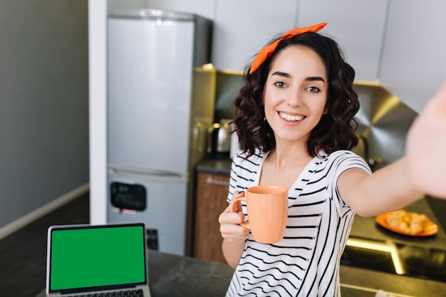 Bonito retrato hermoso selfie de increíble mujer feliz alegre con corte de pelo rizado morena escalofriante en cocina en apartamento moderno. Divirtiéndose, bebiendo té