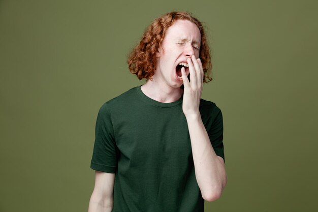 Bostezo boca cubierta con la mano joven guapo con camiseta verde aislado sobre fondo verde