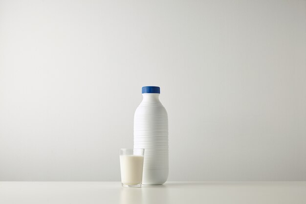 Botella blanca de plástico en blanco con tapa azul cerca del vaso con leche aislado en el centro de la mesa blanca, textura rica