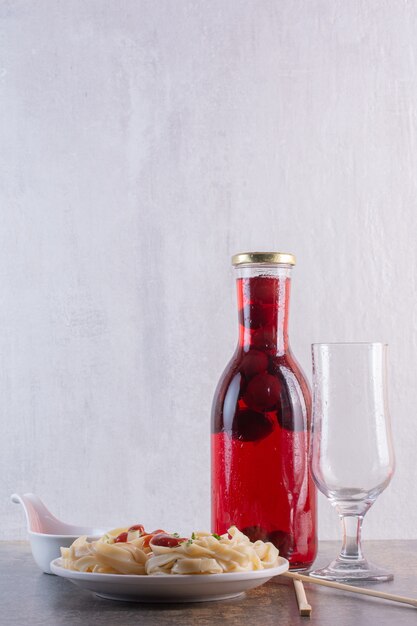 Botella de jugo rojo con vaso vacío y pasta sobre superficie blanca