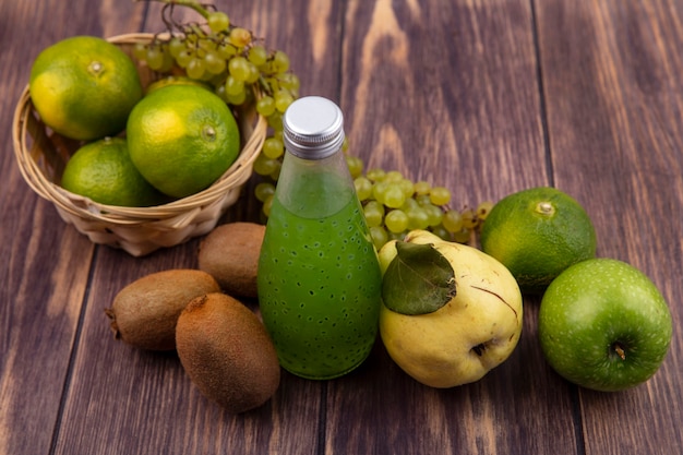 Botella de jugo de vista lateral con pera, kiwi, mandarinas, manzanas y uvas en una canasta en una pared de madera