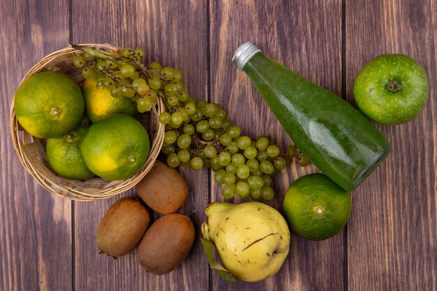 Botella de jugo de vista superior con pera, kiwi, mandarinas, manzanas y uvas en una canasta en una pared de madera