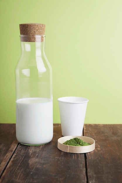 Botella de leche vintage, vidrio para llevar de papel blanco y polvo de té matcha orgánico premium en una mesa de madera cepillada marrón frente a un fondo verde simple