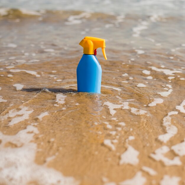 Botella de plástico protector solar azul en el agua de mar poco profunda en la playa