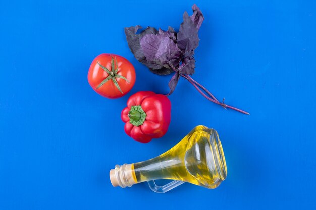 Botella de vidrio de aceite virgen extra con albahaca, pimiento rojo y tomate sobre mesa azul.