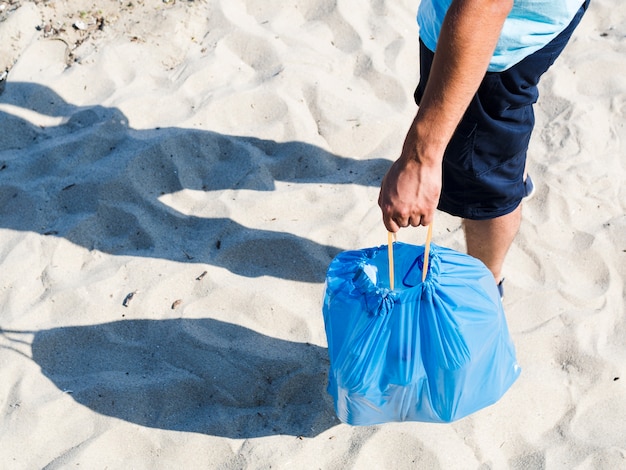 Botellas de plástico en bolsa azul por hombre de pie sobre la arena