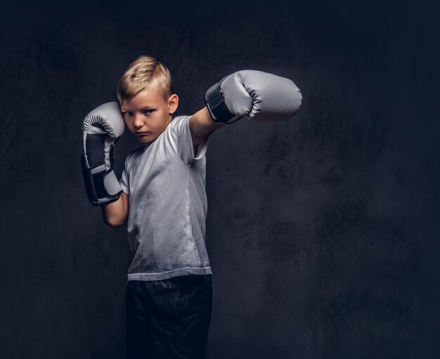 El boxeador escolar con cabello rubio vestido con una camiseta blanca con guantes de boxeo muestra una patada de boxeo. Aislado en el fondo oscuro con textura.