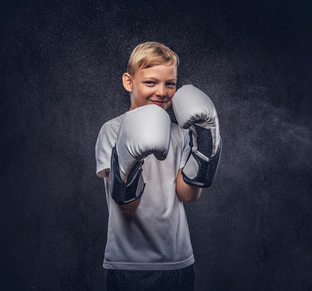 Boxeador joven alegre con cabello rubio vestido con una camiseta blanca con guantes de boxeo posando en un estudio. Aislado en el fondo oscuro con textura.