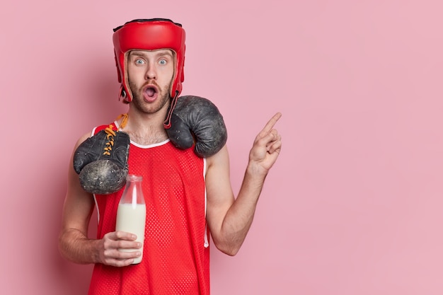 El boxeador masculino activo sorprendido vestido con ropa deportiva tiene guantes de boxeo alrededor del cuello y sostiene una botella de leche como fuente de calcio apunta en el espacio de la copia.