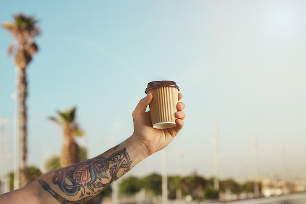 Foto gratuita el brazo y la mano del hombre tatuado con una taza de café desechable beige de cartón ondulado contra el cielo azul claro y palmeras