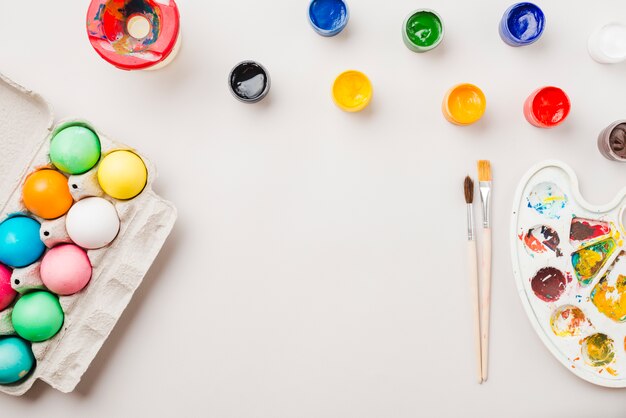 Brillante colección de huevos de colores cerca de un recipiente cerca de pinceles, acuarelas y paleta