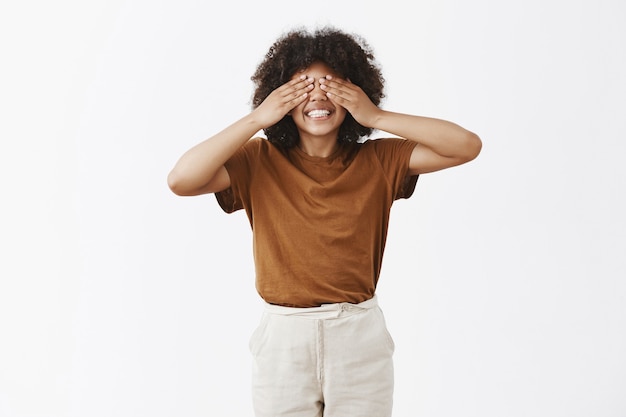 brillante, feliz y joven, atractiva, afroamericana, chica bajita con peinado afro cubriéndose los ojos con las palmas y sonriendo despreocupado como si estuviera jugando a las escondidas estando de humor lúdico