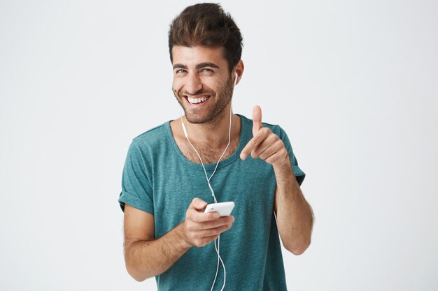 Brillantemente sonriente chico español sin afeitar en camiseta azul, sosteniendo el teléfono inteligente, escuchando música con auriculares, riendo y gesticulando. Expresiones y emociones faciales humanas positivas