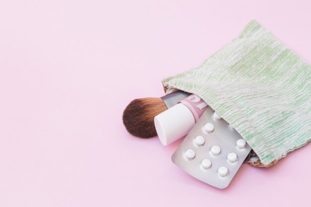 Brocha de maquillaje; Botella de barniz de uñas y blister de pastillas blancas dentro de la bolsa de algodón sobre fondo rosa