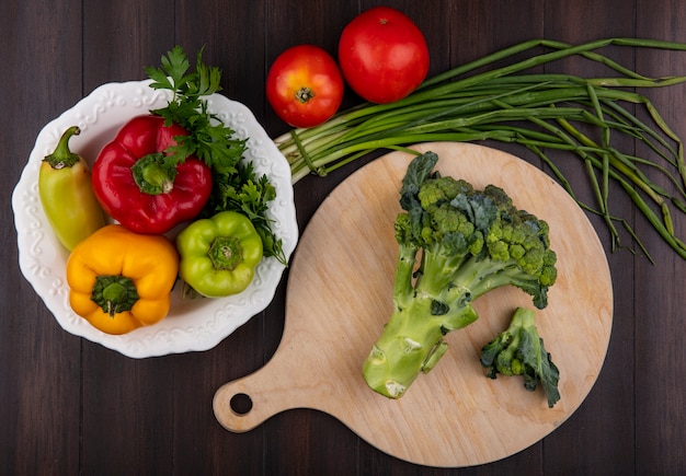 Foto gratuita brócoli de vista superior en una tabla de cortar con pimientos en un plato y cebollas verdes con tomates sobre un fondo de madera