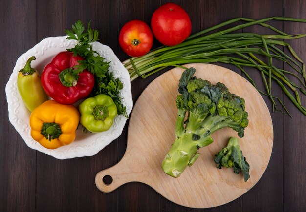 Brócoli de vista superior en una tabla de cortar con pimientos en un plato y cebollas verdes con tomates sobre un fondo de madera