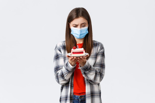 Brote de coronavirus, estilo de vida durante el distanciamiento social y concepto de celebración navideña. Mujer confundida en máscara médica mirando perplejo la vela encendida de la torta de cumpleaños, pensando, fondo blanco.