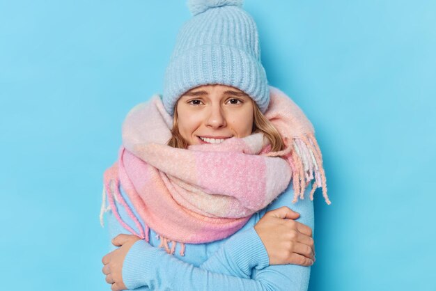 Brr qué helada allí. Mujer joven se abraza y se siente frío pide encender el calentador usa gorro de punto bufanda caliente alrededor del cuello camina durante el frío día de invierno escalofríos aislados sobre fondo azul