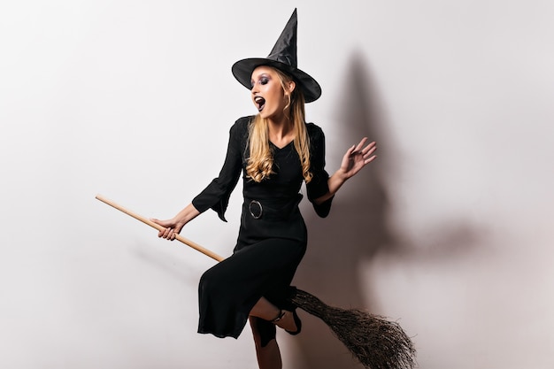 Bruja alegre volando en escoba en halloween. Retrato de interior de maga entusiasta en vestido negro.