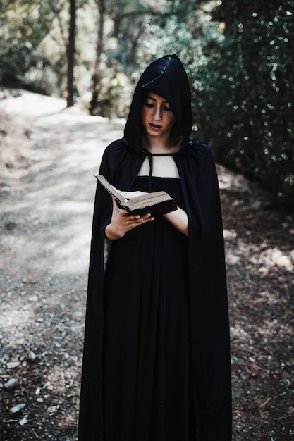 Bruja encapuchada con el libro en el bosque durante el día