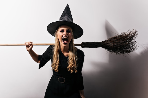 Bruja enojada con el pelo largo con escoba. Asistente mujer rubia gritando en halloween.