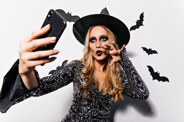 Bruja entusiasta con maquillaje oscuro haciendo selfie con murciélagos. vampiro femenino glamoroso posando en la pared blanca.