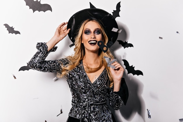 Bruja glamorosa emocionada con maquillaje negro riendo. Vampiro rubio sonriente con sombrero relajante en halloween.