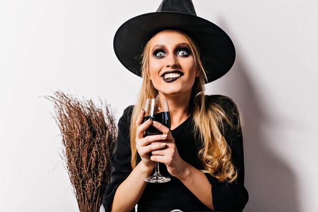 Bruja loca bebiendo sangre en la pared blanca. espectacular maga sosteniendo copa de vino con poción.