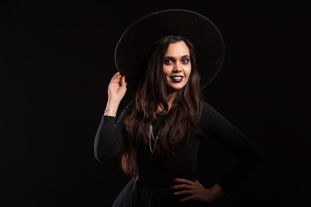 Bruja morena joven hermosa que sostiene su sombrero con su mano sobre fondo negro. Maquillaje para halloween. Mujer bonita vestida de bruja para halloween.