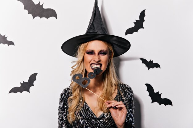 Bruja con peinado ondulado celebrando halloween. Vampiro mujer extática con sombrero divertido posando con murciélagos.