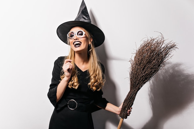 Bruja rizada en vasos que expresan felicidad en halloween. Foto interior de niña bonita riendo en traje de mago sosteniendo una escoba.